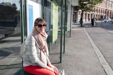 Bahar güneşli bir günde otobüs durağı üzerinde oturan şık hippi kız. şık kadın turist şehir ulaşım için bekliyor