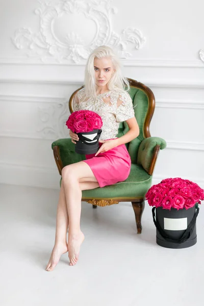年轻漂亮的女孩与可爱的脸和长金色的头发。女人坐在那里, 拿着粉红色玫瑰的黑盒子。豪华内饰 — 图库照片