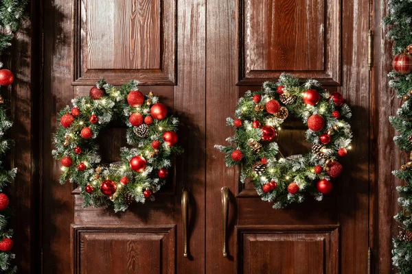 Είσοδος σπιτιού διακοσμημένη για διακοπές. Χριστουγεννιάτικη διακόσμηση. Δύο στεφάνια και γιρλάντα από κλαδιά ελάτης. Μεγάλη ξύλινη πόρτα — Φωτογραφία Αρχείου