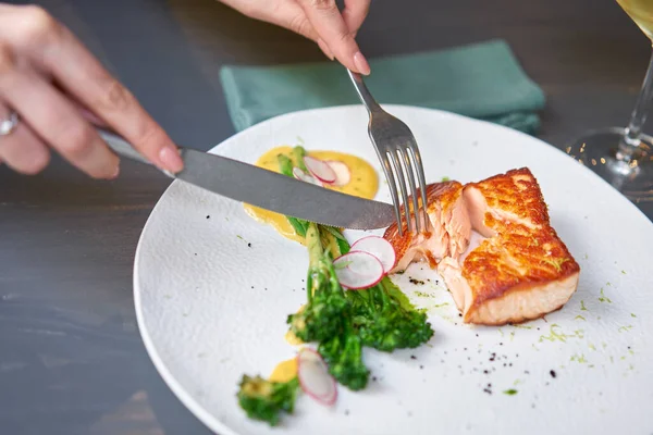 Lunch in a restaurant, a woman cuts the Salmon steak fillet and garnished with young broccoli. Restaurant menu, a series of photos of different dishes — Stock Photo, Image