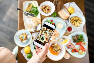 Bir blogcu sosyal ağlar için akıllı telefondan fotoğraf çekiyor. Süslü deniz ürünleri sandviçi tabağı. Tabakların çeşitleri masada. Restoran menüsü.