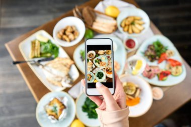 Bir blogcu sosyal ağlar için akıllı telefondan fotoğraf çekiyor. Süslü deniz ürünleri sandviçi tabağı. Tabakların çeşitleri masada. Restoran menüsü.