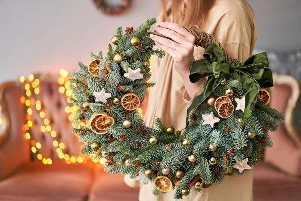 Christmas mood. Beautiful festive wreath of fresh spruce in woman hands. Xmas tree. Bokeh of Garland lights on background.
