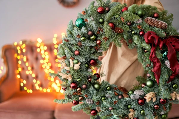 Χριστουγεννιάτικη διάθεση. Όμορφο εορταστικό στεφάνι από φρέσκο έλατο σε γυναικεία χέρια. Χριστουγεννιάτικο δέντρο. Bokeh του Garland φώτα στο παρασκήνιο. — Φωτογραφία Αρχείου