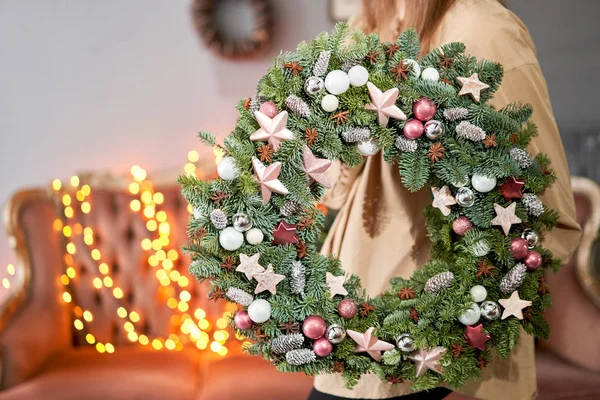 Kerststemming. Prachtige feestelijke krans van verse sparren in vrouwenhanden. Kerstboom. Bokeh van Garland lichten op de achtergrond. — Stockfoto