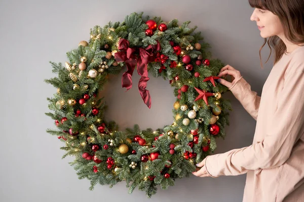 Prachtige feestelijke krans van verse sparren in vrouwenhanden. Kerst cirkel met rode en gouden ornamenten en ballen. Kerststemming. Grijze muur op achtergrond. — Stockfoto