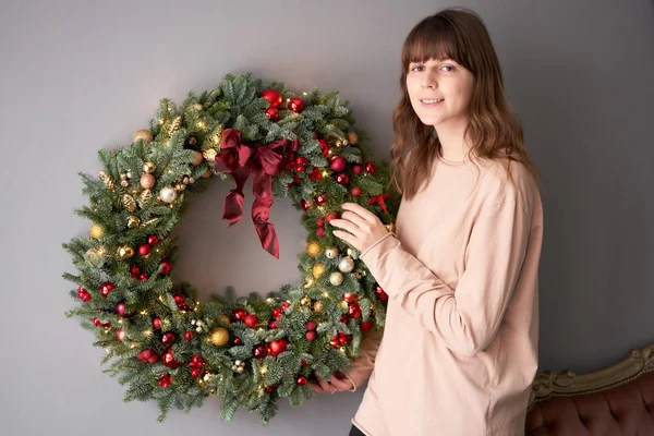 Prachtige feestelijke krans van verse sparren in vrouwenhanden. Kerst cirkel met rode en gouden ornamenten en ballen. Kerststemming. Grijze muur op achtergrond. — Stockfoto