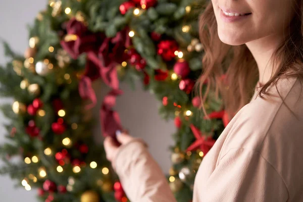 Prachtige feestelijke krans van verse sparren in vrouwenhanden. Kerst cirkel met rode en gouden ornamenten en ballen. Kerststemming. Grijze muur op achtergrond. — Stockfoto