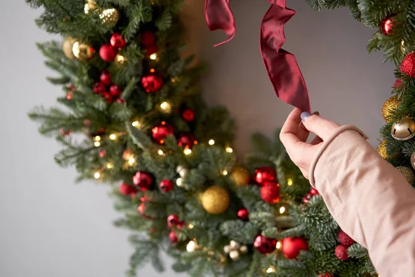 Bella corona festiva di abete rosso fresco nelle mani della donna. Cerchietto natalizio con ornamenti e palline in oro e rosso. L'umore natalizio. Parete grigia su sfondo . — Foto Stock