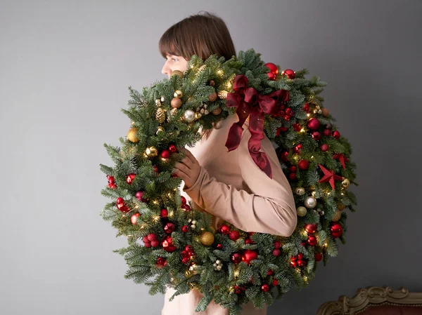 Bela grinalda festiva de abeto fresco em mãos de mulher. Xmas circlet com ornamentos vermelhos e dourados e bolas. Humor de Natal. Parede cinza no fundo . — Fotografia de Stock