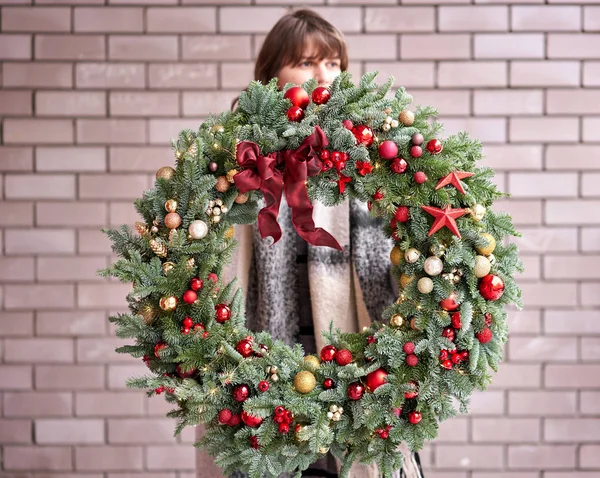 Beautiful festive wreath of fresh spruce in woman hands. Xmas circlet with red and gold ornaments and balls. Christmas mood. Brick wall on background.