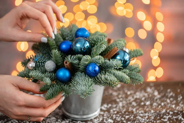 Vrouw versiert kleine arrangement van verse sparren in een metalen pot. Kerststemming. Garland Bokeh op de achtergrond. Bokeh van Garland lichten op de achtergrond — Stockfoto