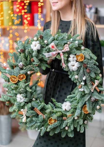 Hermosa corona festiva de abeto fresco en manos de mujer. Humor de Navidad. Bokeh de luces de guirnalda en el fondo . — Foto de Stock