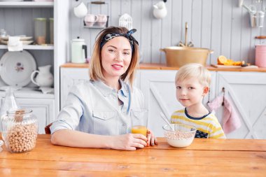 Anne ve oğlu mutfakta kahvaltı ederken gülümsüyorlar. Mutfakta aydınlık bir sabah. Sağlıklı kahvaltı gevrekleri ve taze meyve..