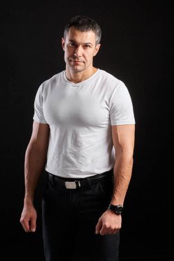 Beyaz tişörtlü yakışıklı atletik adam siyah duvar arkasında duruyor.