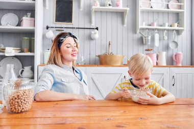 Çocuk mısır gevreğini sütle yiyor. Anne ve oğlu mutfakta kahvaltı ederken gülümsüyorlar. Mutfakta aydınlık bir sabah. Sağlıklı kahvaltı gevrekleri ve taze meyve..