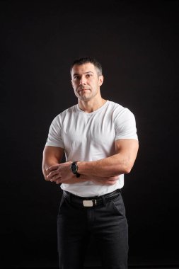 Beyaz tişörtlü yakışıklı atletik adam siyah duvar arkasında duruyor.