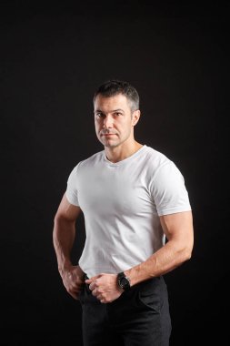 Beyaz tişörtlü yakışıklı atletik adam siyah duvar arkasında duruyor.