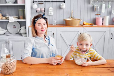 Çocuk aç gözlülükle mısır gevreğini sütle yiyor. Anne ve oğlu mutfakta kahvaltı ederken gülümsüyorlar. Mutfakta aydınlık bir sabah. Sağlıklı kahvaltı gevrekleri ve taze meyve..
