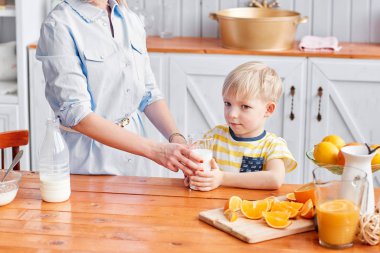 Çocuk bir bardaktan süt içer. Anne ve oğlu mutfakta kahvaltı ederken gülümsüyorlar. Annem bardağa süt döküyor.
