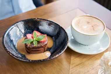 Kadın çikolatalı kek yiyor, karamelli, çilek ve nane yaprağıyla süslenmiş. Kafede kahvaltı, sabah kahvesi. Tahta masada. Restoran menüsü