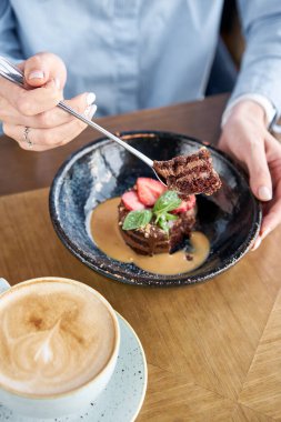 Kadın çikolatalı kek yiyor, karamelli, çilek ve nane yaprağıyla süslenmiş. Kafede kahvaltı, sabah kahvesi. Tahta masada. Restoran menüsü