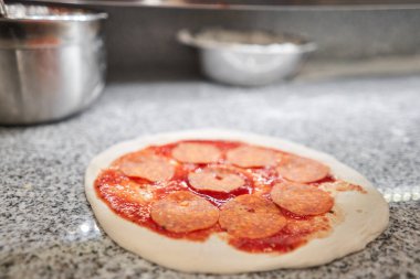 Pizza pişiriyorum. Peynir ve salam doldurur, hamur preformundaki malzemeler. Mutfakta üniformalı beyaz önlüklü aşçının yakın çekimi.