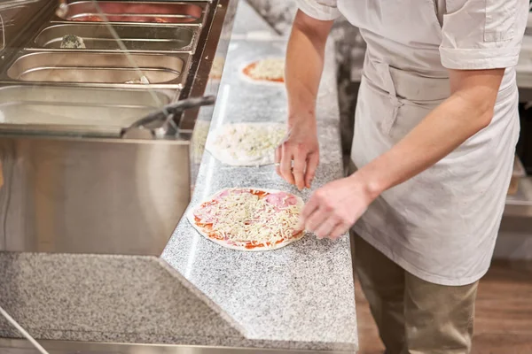 Μαγειρεύω πίτσα. οργανώνει τα συστατικά τυριού στο πρόπλασμα ζύμης. Κοντινό χέρι του σεφ φούρναρη με στολή λευκό ποδιά μάγειρας στην κουζίνα — Φωτογραφία Αρχείου