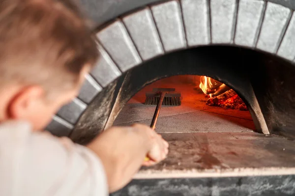 ピザシェフは特別なブラシでピザオーブンを掃除します。イタリアのレストランで伝統的な木のオーブン。オリジナルのナポリのピザ。赤熱石炭. ストック画像