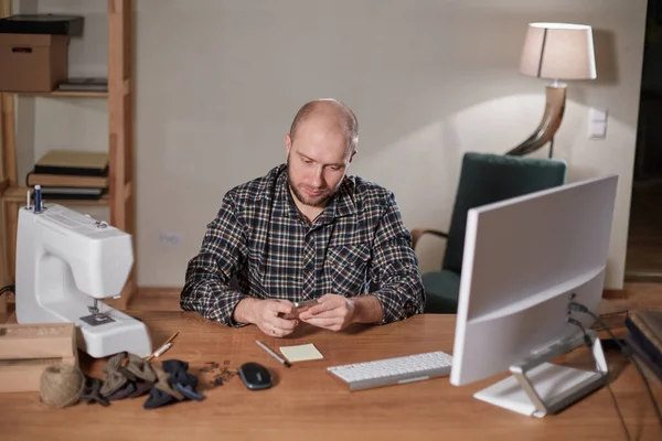 Mannen på datorn ser ut leveransen av webbutiken. Arbetar med symaskin i textilateljé. Skräddarsydda slipsar av ulltyg. — Stockfoto