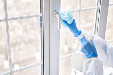 Dezenfeksiyon, temizlik ve pencerenin kulpunu yıkama. Koruyucu takım elbiseli ve mavi eldivenli bir kadın dezenfektan solüsyonu sıkar. Koronavirüs enfeksiyonunun önlenmesi.