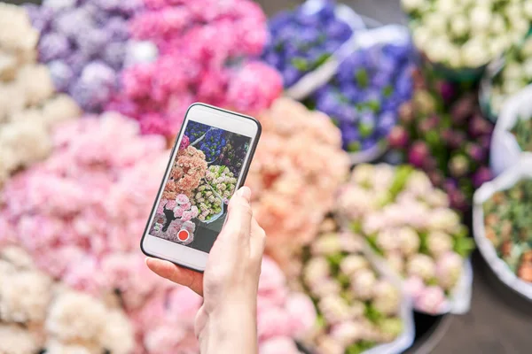 Entrega de flores. Una florista mujer toma una foto en su teléfono de flores. Concepto de tienda floral. Florista mujer crea arreglo floral en una canasta de mimbre . — Foto de Stock