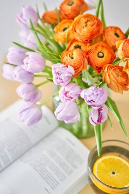Bahar kompozisyonu, leylak laleleri, turuncu düğün çiçekleri ve limonlu bir fincan yeşil çay, ahşap bir masada açık bir kitap. Ev konseptinde kal