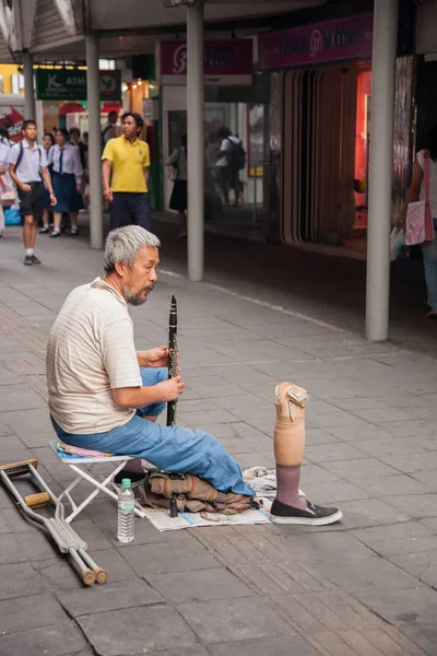 Um deficiente toca clarinete na calçada, em uma rua de Bangkok com uma perna artificial na frente dele . — Fotografia de Stock