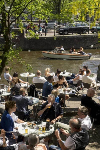 Los turistas toman una copa al sol en la terraza de un café a orillas de un canal en Ámsterdam — Foto de Stock