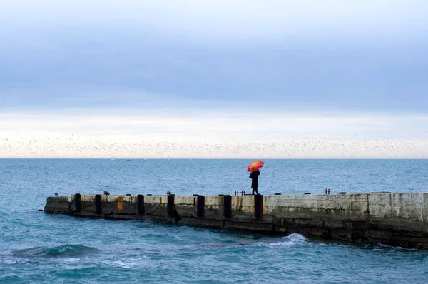明るい傘を持った女性が海のほとりの桟橋を歩いている ストックフォト
