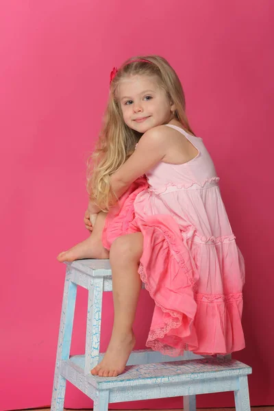Маленька дівчинка в одязі сидить на стільці. Рожевий фон — стокове фото