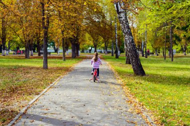 Parlak bir sonbahar parkında güzel bir manzara. Yolda bisiklete binen küçük kız.