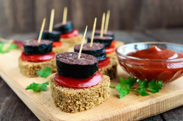 Сэндвичи с черным ржаным хлебом в форме сердца, кровяная колбаса (Morcillo) и кусочки сладкого перца на шампуре, томатный соус на темном деревянном фоне . — стоковое фото
