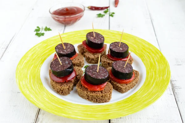 Sandwiches mit Schwarzbrot in Herzform, Blutwurst (Morcillo) und Paprikastücken auf Spießen und Tomatensauce, auf weißem Holzgrund. — Stockfoto