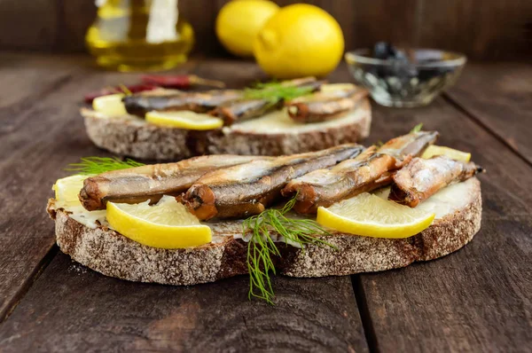 Sandwich met gerookte vis lodde (sprotten), citroen op Zwarte roggebrood. Snack op een donkere houten tafel. — Stockfoto