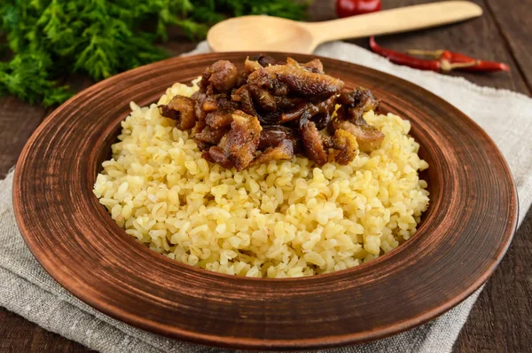 Eastern traditional wheat porridge - bulgur with roasted pieces fatty meats, bacon in a clay bowl on dark wooden background. — Stock Photo, Image