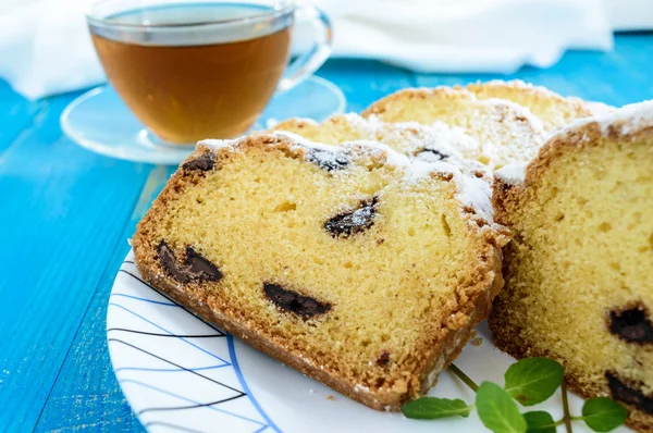 Φρεσκοψημένο σπιτικό κέικ, διακόσμηση από την κορυφή με ζάχαρη άχνη, κομμένα φέτες, με κομμάτια σοκολάτας σε ένα πιάτο και φλιτζάνι τσάι σε μπλε φόντο. Κοντινό πλάνο — Φωτογραφία Αρχείου