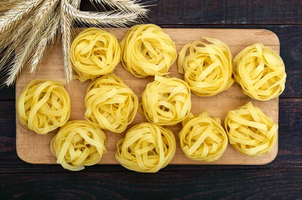 Pasta tagliatelle in the form of nests on a cutting board on a dark wooden background with spikelets of wheat. The top view. — Stock Photo, Image