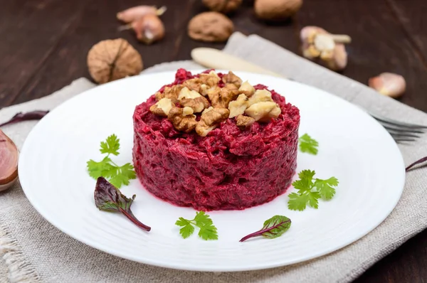 Vegetarian Lenten dish: a salad of beets with walnuts and garlic in a white plate in the shape of a circle on a dark wooden background. — Stock Photo, Image