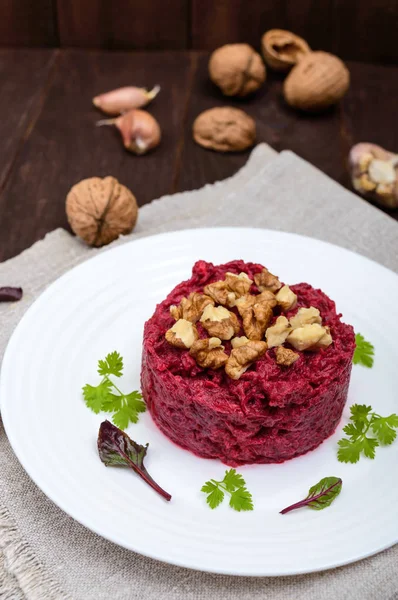 Vegetarian Lenten dish: a salad of beets with walnuts and garlic in a white plate in the shape of a circle on a dark wooden background. Vertical view — Stock Photo, Image