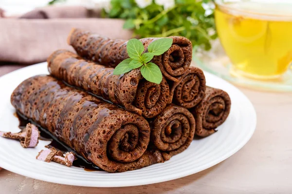 Dünne, zarte Schokoladenkuchen, gerollt, in einem Haufen auf einem weißen Teller und einer Tasse Kräutertee mit Honig ausgebreitet — Stockfoto