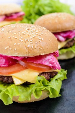 Hamburger sulu pirzola, domates, lahana turşusu ve salatalık, peynir, yeşil marul yaprakları ve siyah arka plan üzerine susam ile yumuşak bir topuz ile