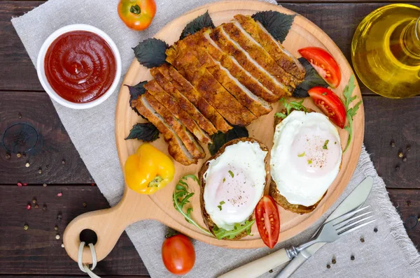 Trozos de chuleta (schnitzel), tostadas con huevos, tomate fresco sobre una tabla de madera sobre un fondo oscuro. Vista superior. — Foto de Stock
