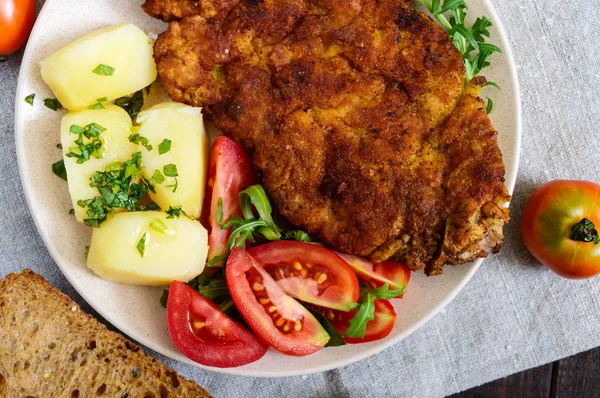 Altın şnitzel, haşlanmış patates ve koyu ahşap arka plan üzerinde bir tabakta domates salatası.